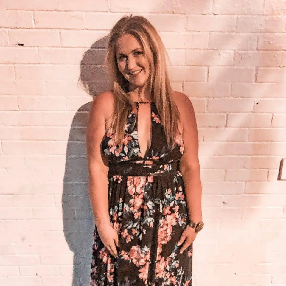 Woman in floral dress standing against brick wall.